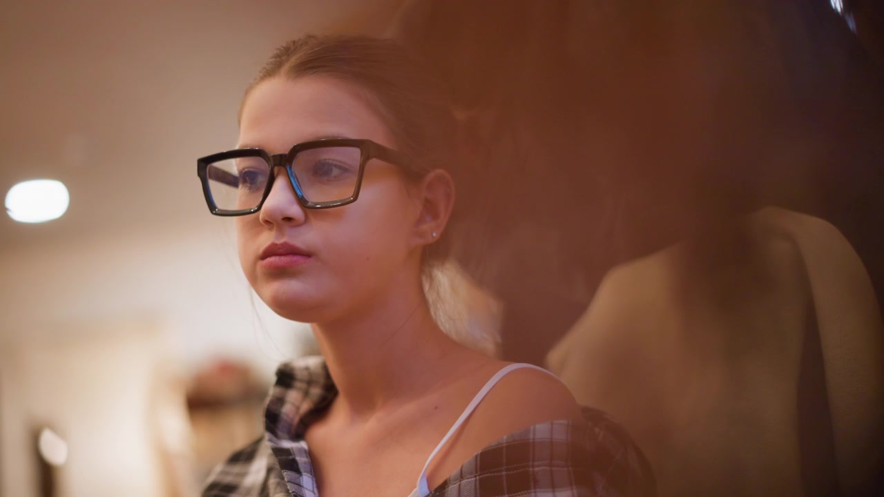 Primo piano di giovane donna con occhiali e camicia a quadri, delicatamente illuminata in un ambiente interno caldo. La donna rimane immobile indossando abiti casual, incluse le spalline visibili della canotta sotto il colletto aperto