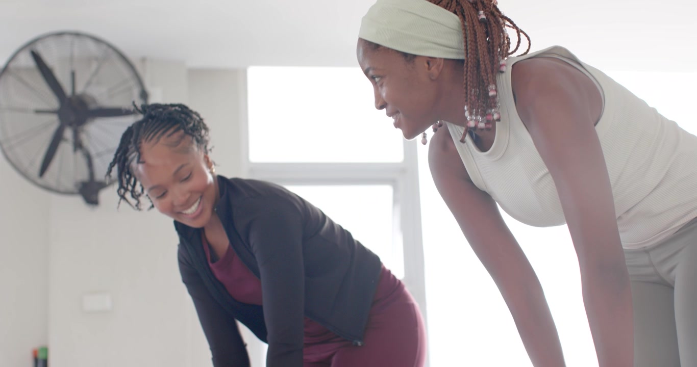 African female friends wearing leggings and tanks, deepening fold with blue bands under feet in gym