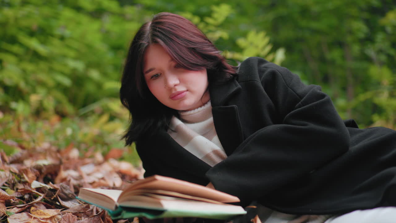 Stylish lady lies on leaf carpet in forest, focused on open book, flipping pages attentively, cozy black coat, soft green background, calm autumn study moment, quiet outdoor reading scene