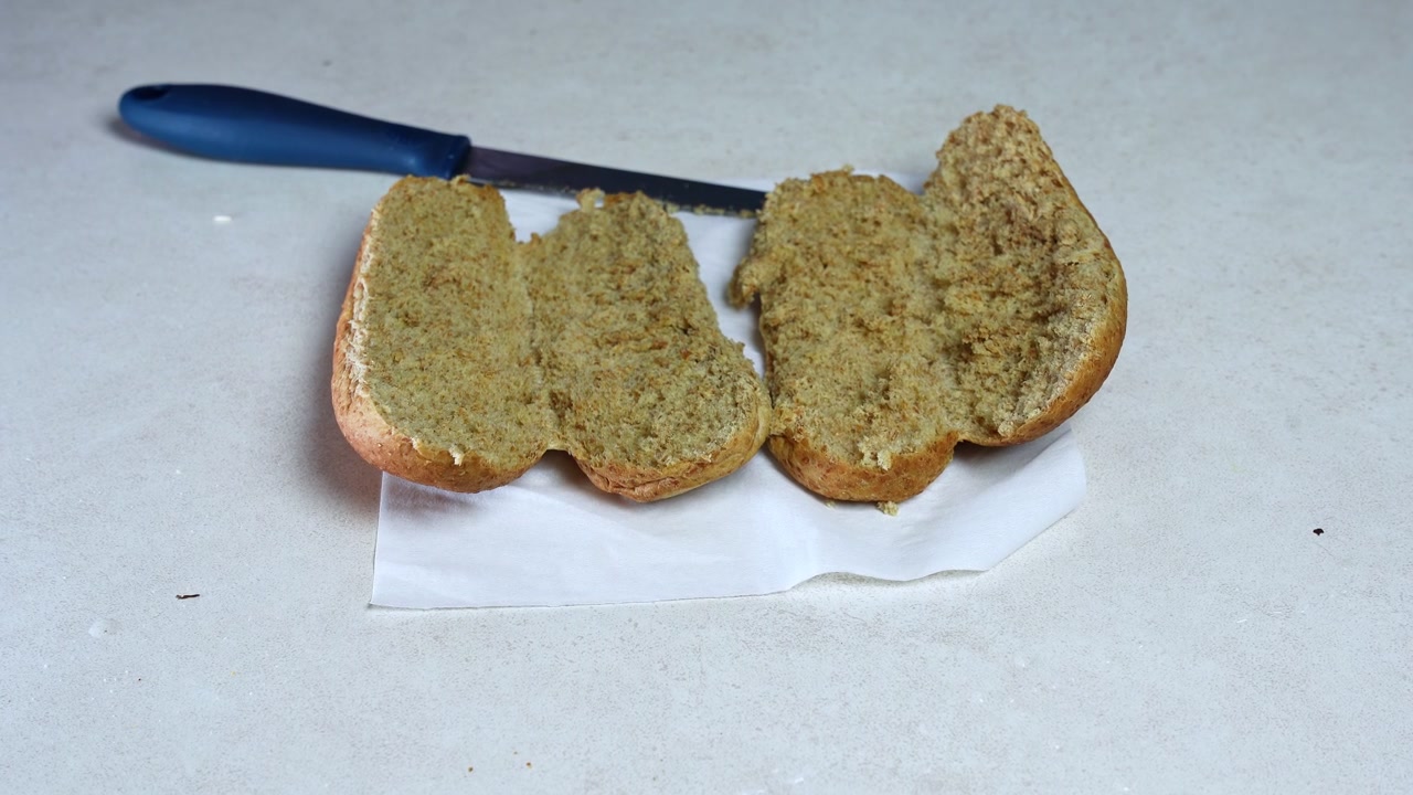 A close-up static shot of a freshly sliced brown whole wheat bread roll resting on a white paper napkin. A blue-handled kitchen knife lies next to the food on a light stone indoor kitchen counter.
