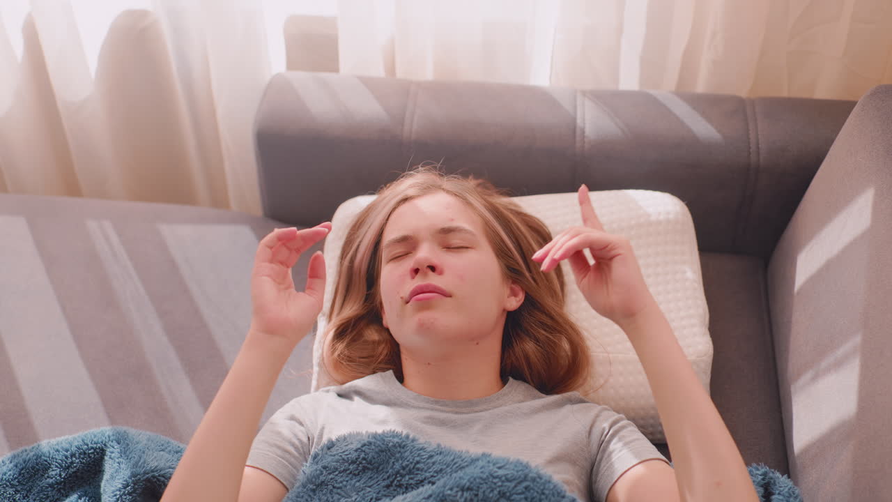 Top down view of lady dancing gently on couch covered in blanket, enjoying peaceful morning relaxation with eyes closed, sunlight streaming through curtains
