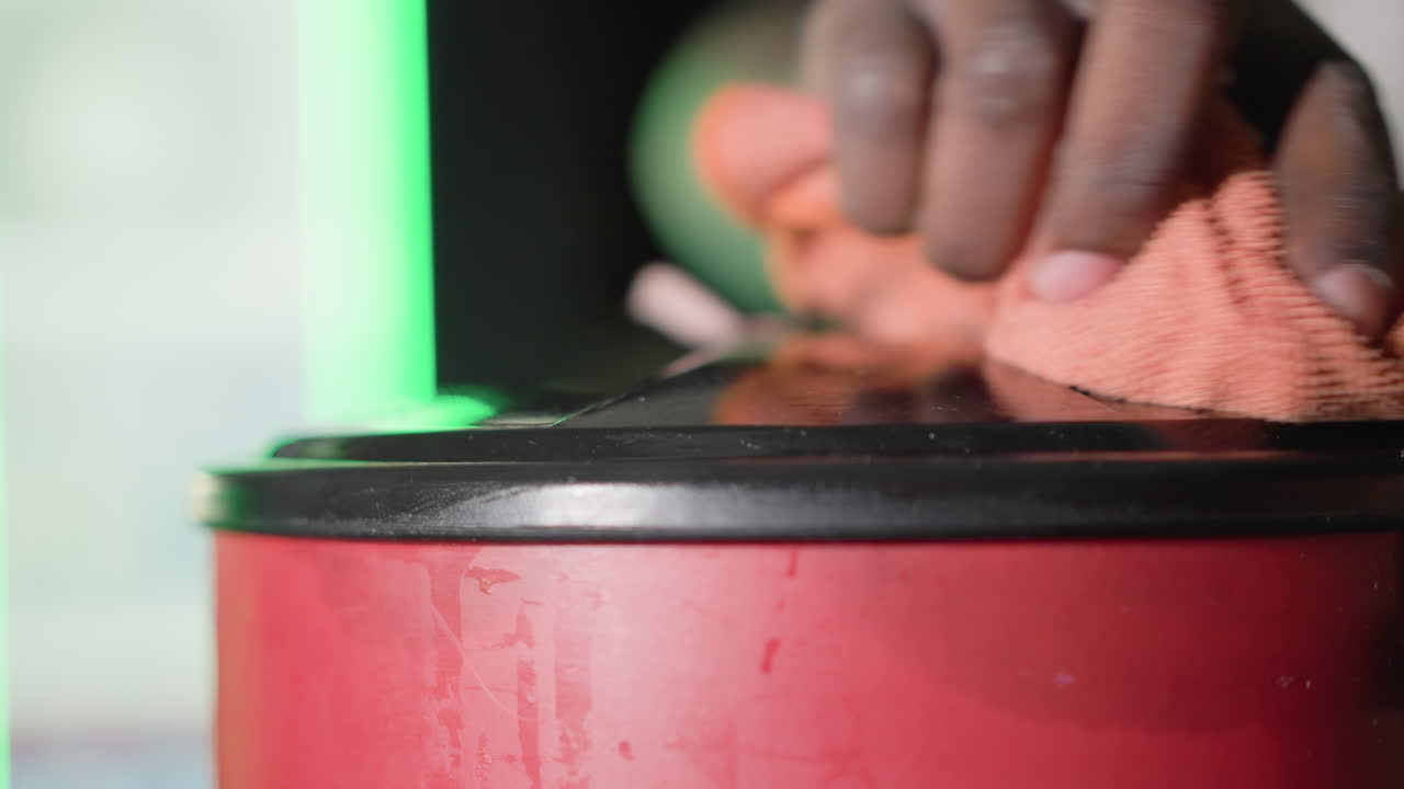 Close-up shot of bartender cleaning red bin with orange cloth, Heineken neon green light glowing in background. Bartender wears white t-shirt, focused on cleaning task behind bar