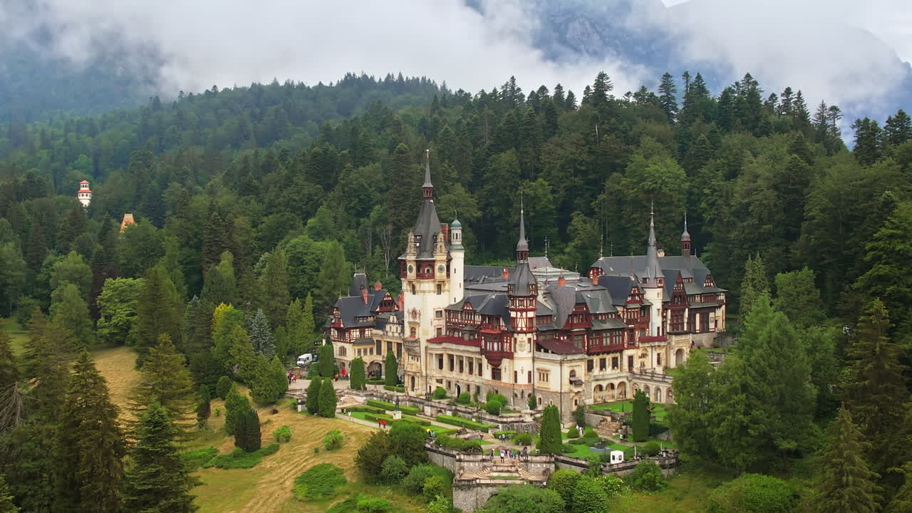 Luftfoto med drone af Peleș-slottet i Rumænien. Slot med haver og turister i Karpaterne, frodig skov omkring.