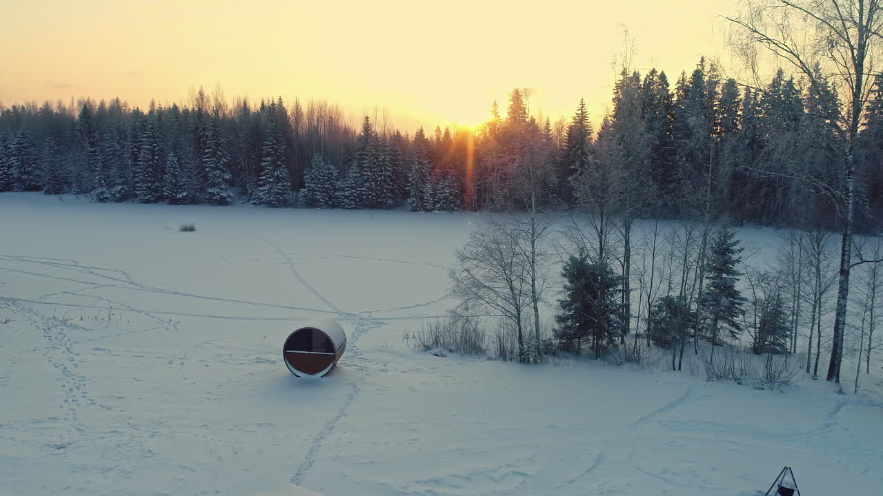 un dron aéreo se disparó hacia atrás sobre una cabaña rectangular y una sauna de barril cubierta de nieve blanca a lo largo del paisaje rural durante la noche