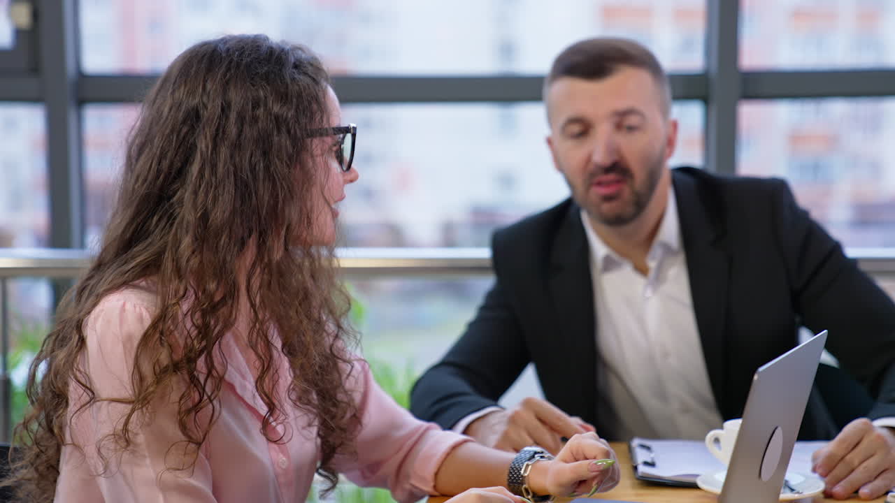 Office team members sit at the table and brainstorming at cup of coffee. Male boss addresses female employee and both look at computer.
