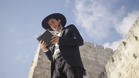 pan shot of an orthodox male in a black suit and hat, praying and reading from a tehilim book beside an ancient old city in jerusalem