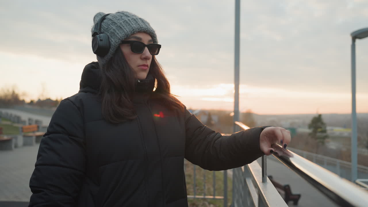 Sul tetto al tramonto, una persona in cappotto vicino alla ringhiera metallica, con berretto di lana e occhiali da sole, compie un lento gesto della mano simile a una danza. Skyline crepuscolare cinematografico, umore introspettivo. Un pendolare urbano divenuto performer/modello.