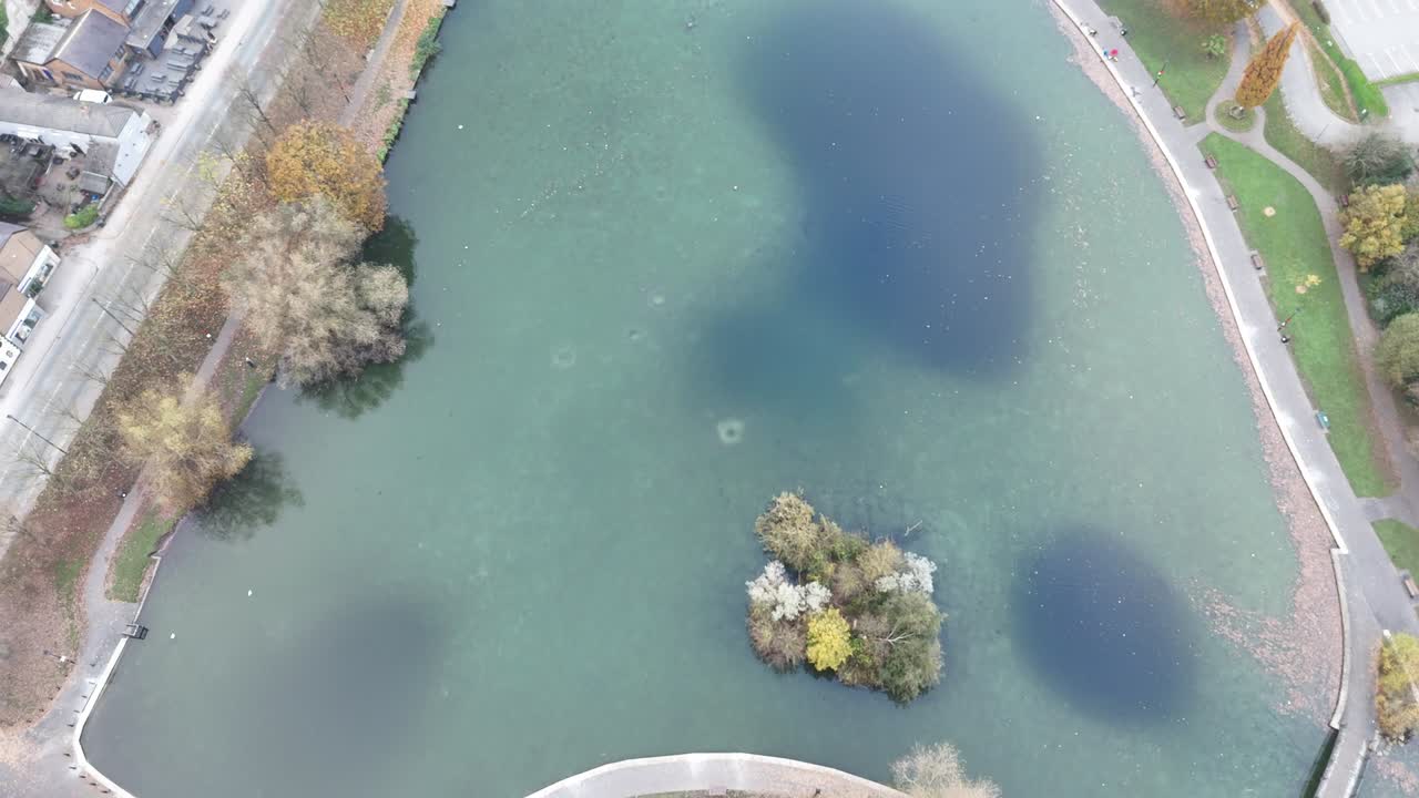 Drone top view of clear pond with small island of trees in center, surrounded by curved path, road and buildings. Autumn trees add color to peaceful urban landscape in Bochum