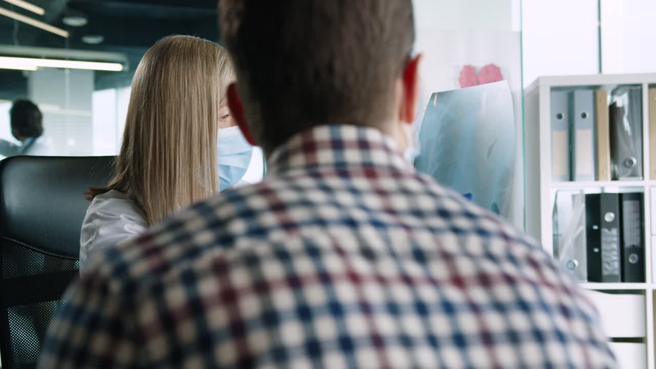 의료 상담에서 남성 환자에게 코로나 바이러스 폐 질환을 설명하는 책상에 앉아 의료 마스크를 착용하는 백인 여성 의사