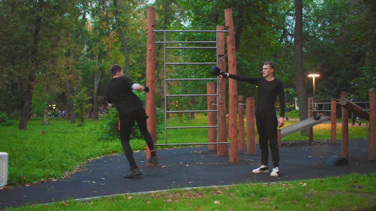 outdoor park training two men practicing kicks by wooden calisthenics bars at dusk, partner holds mitts while other performs sidekick, playful energy, casual black clothing, grassy surroundings, lamp