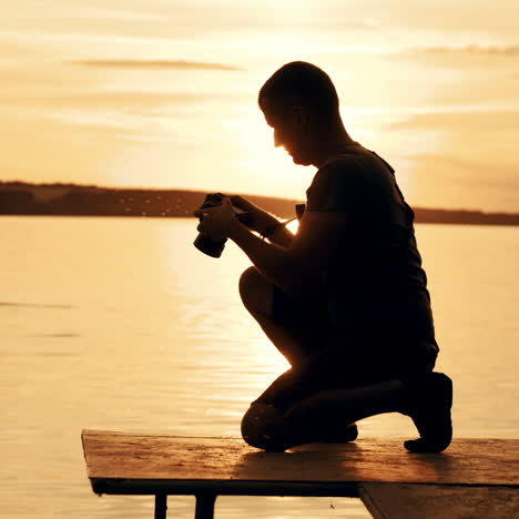 a man in shorts and a t-shirt is squatting and holding a camera focused on the sunset against the background of the forest and people in the river.