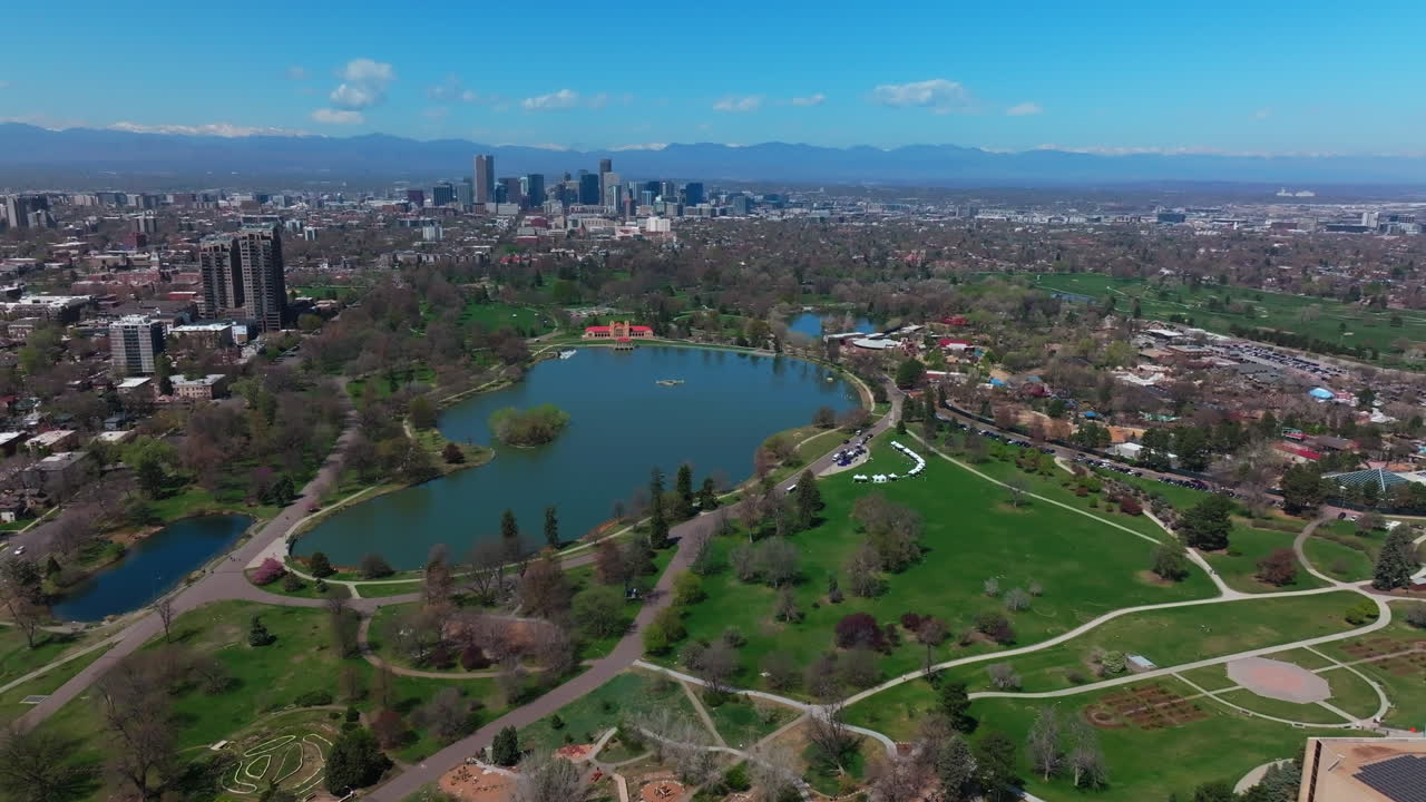 City Park Denver Colorado Lake Pavilion vibrant spring summer aerial drone sunny blue sky snow capped Rocky Mountains front range cityscape green lush grass trees blossom pan left motion