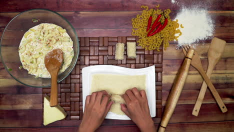 top view of rolling the spring rolls, marinated macaroni in the bowl, raw macaroni, wooden spoon, wooden spatula, flour, rolling pin, and chili on the table