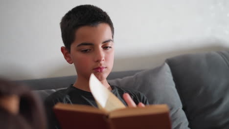 calm boy deeply focused reading on couch flipping through book pages showing intense concentration during indoor study moment with slight view of girl hair beside him