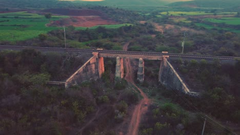 Luftaufnahme-Einer-Alten-Eisenbahnbrücke-Aus-Beton-Mit-Gleisen-Und-Dichten-Waldhügeln-Im-Hintergrund-Während-Der-Späten-Abendzeit-Per-Drohne