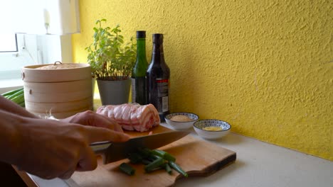 manos masculinas cortando cebollas verdes junto al vientre de cerdo enrollado en la cocina casera