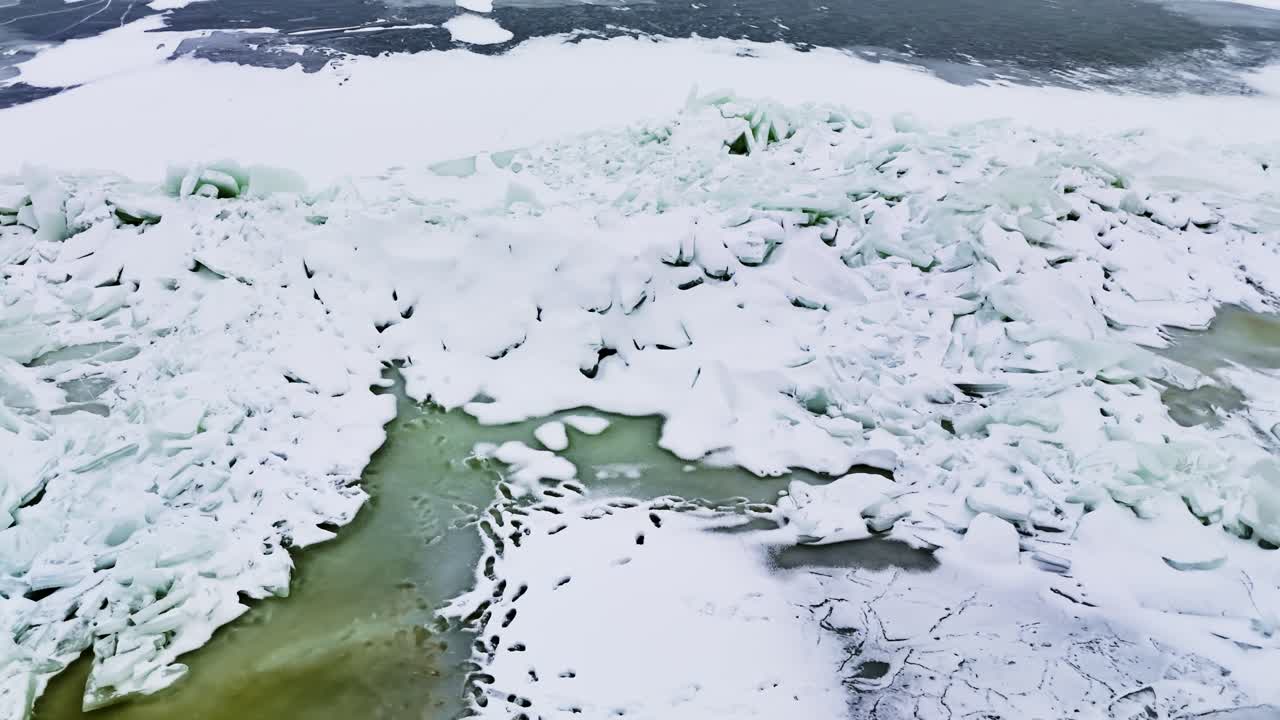 Drone che sorvola una cresta di pressione di ghiaccio marino frantumato e canali di acqua di fusione turchesi lungo la costa del Mar Baltico in Lettonia in una grigia giornata invernale.