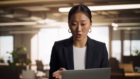 a woman in a blazer intently works on her laptop in a contemporary office. the bustling atmosphere features colleagues engaged in various activities, highlighting a dynamic work environment.