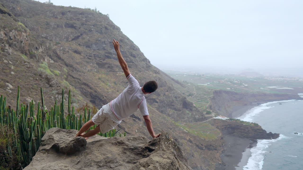 en las montañas, un hombre se equilibra en una mano con la espalda a la cámara, mirando hacia el océano, inmerso en la meditación