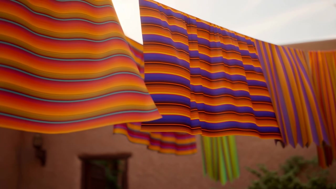 File di tessuti a strisce multicolori appesi a stendibiancheria attraverso un cortile in adobe recintato, visti da un'angolazione bassa con una luce calda e diffusa che evidenzia la trama, il colore e il motivo a strati.