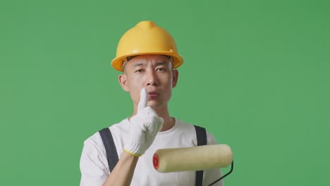 Close Up Of Asian Man Painter Wearing Safety Helmet Looking At Camera And Making Shh Gesture While Standing In The Green Screen Background Studio