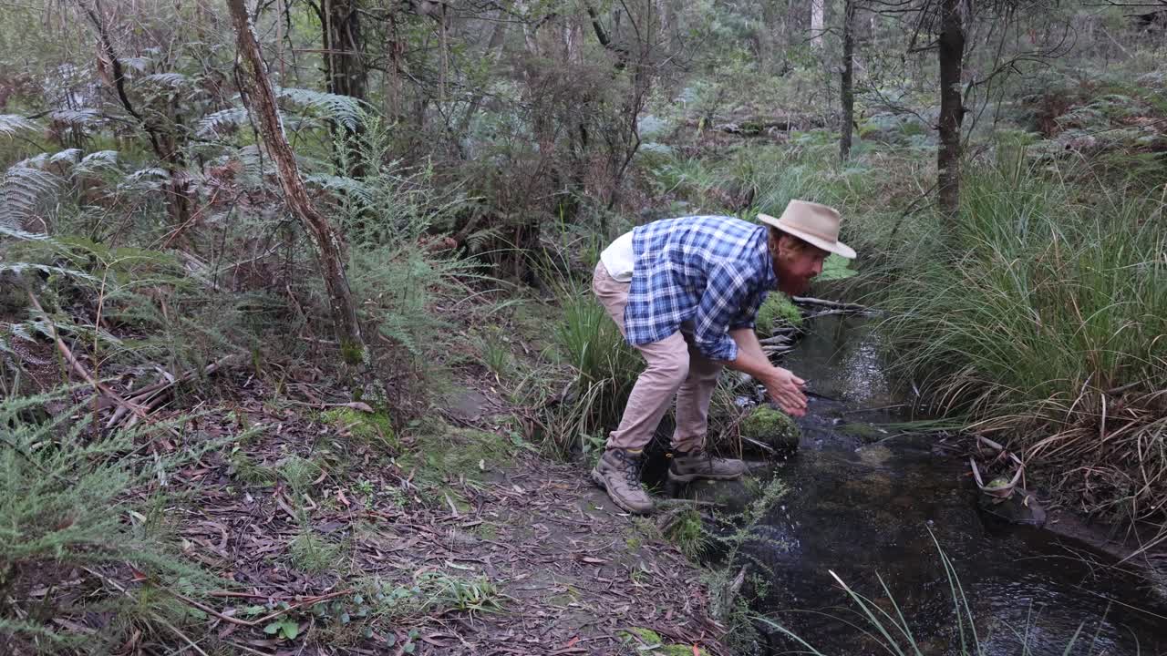 un bosquimano con un sombrero akubra bebe agua dulce de un arroyo en el monte australiano