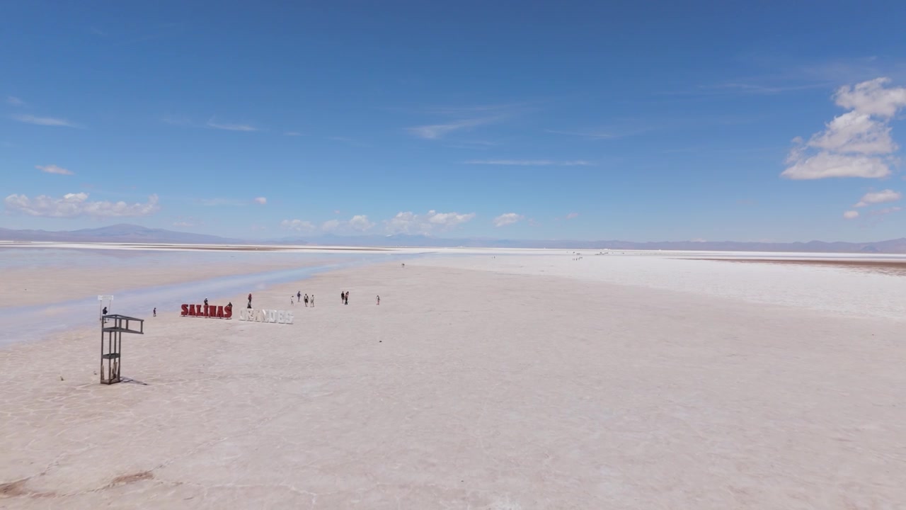 Visitors walking across Salinas Grandes salt flats under clear sky, aerial flyover as they approach welcome sign to tourist location