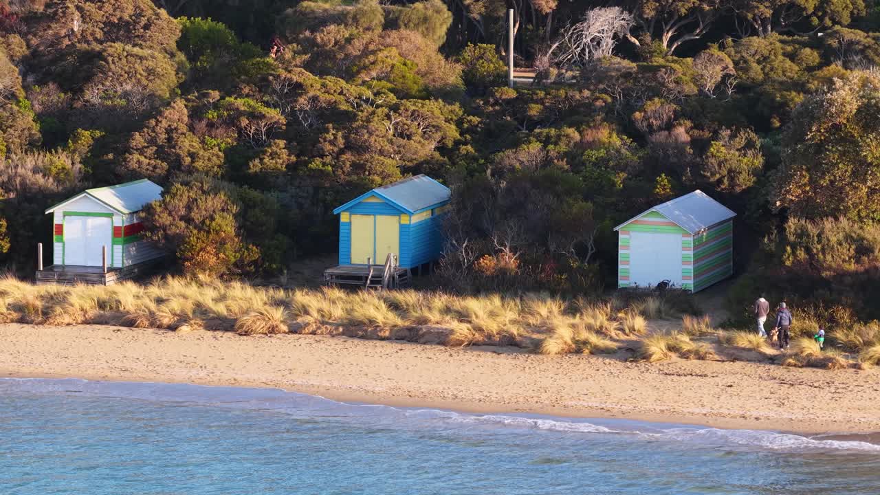 Drone pans over vibrant beach huts, sandy shore, and coastal vegetation in soft afternoon sunlight