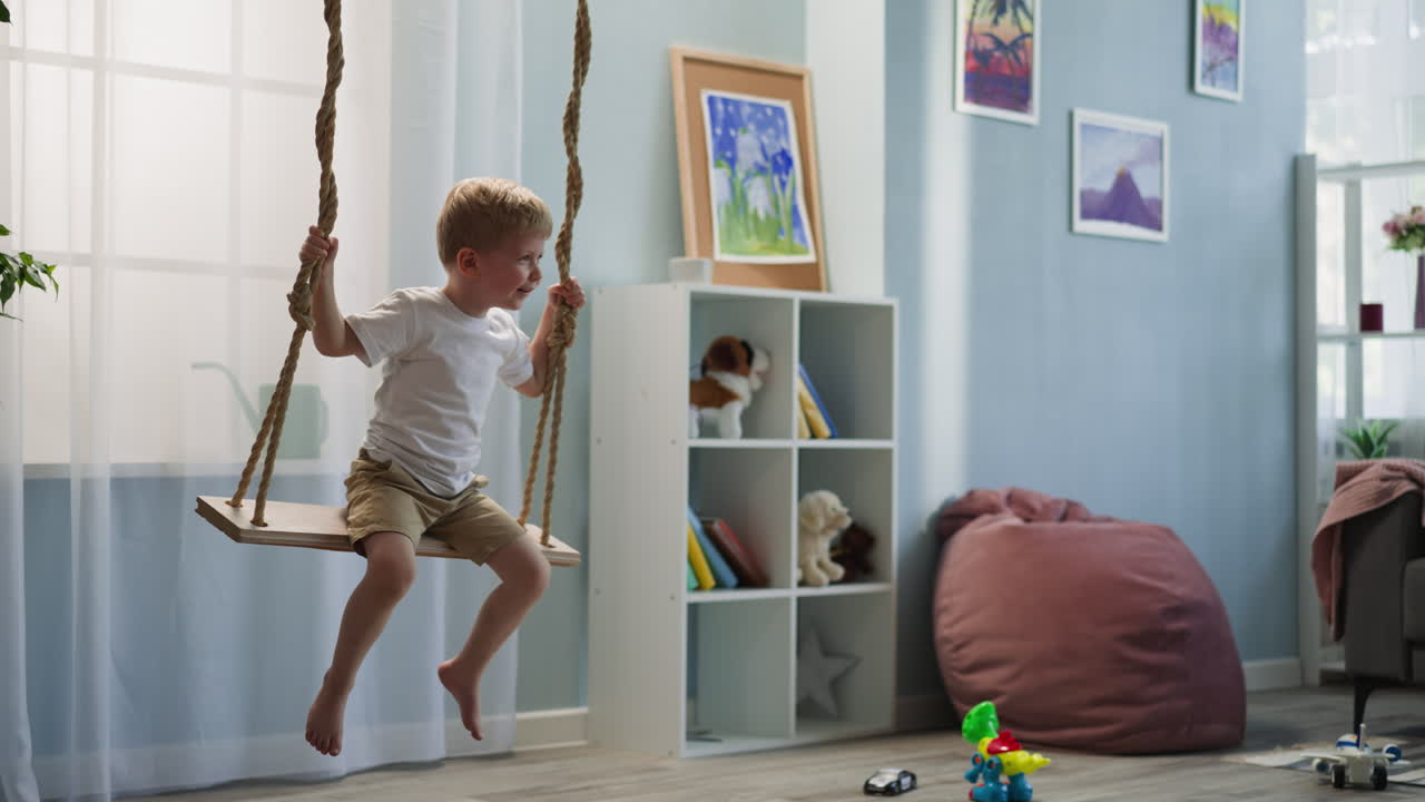 Cheerful little boy sits on wooden swing in room with toys
