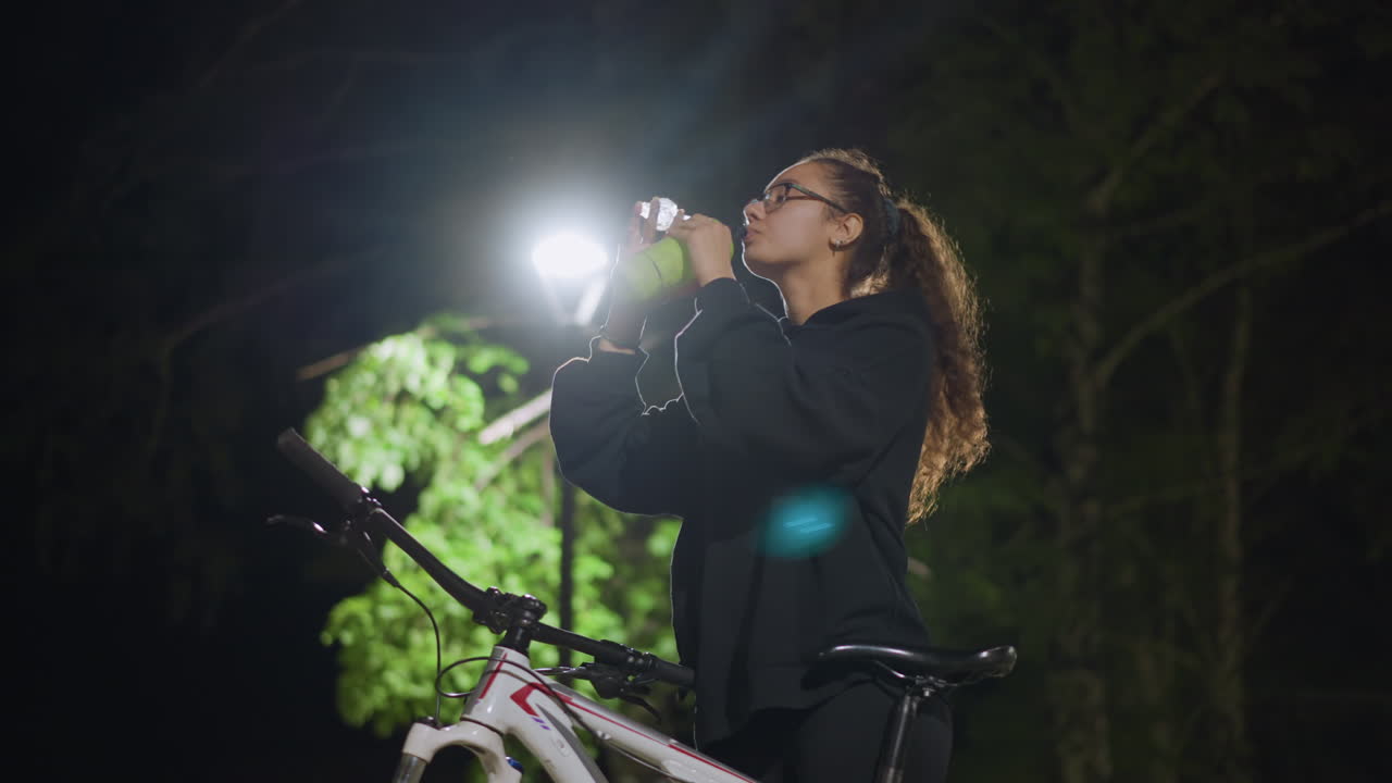 Individual Inspects Pathway Before Evening Ride, Nighttime Cyclist Examines Route Amidst Illuminated Surroundings, Female Cyclist Reviews Route Plan While Adjusting Bottle Under Bright Streetlamps