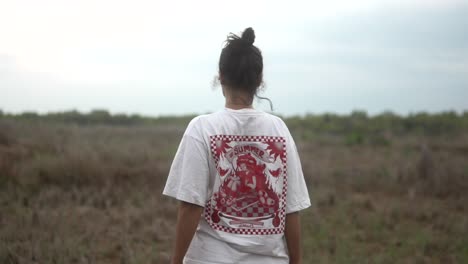 a woman stands in a field wearing a graphic t-shirt, facing away