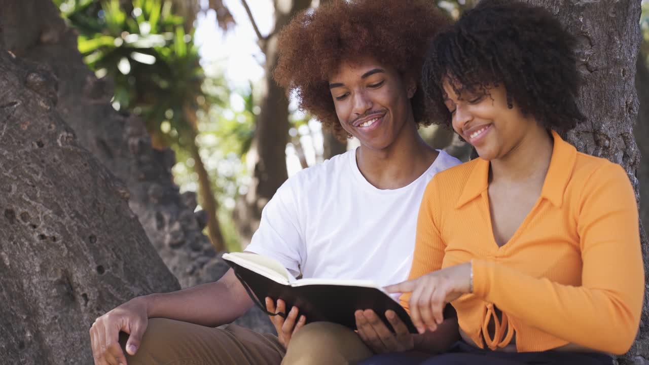 Casal negro feliz sentado lendo um livro perto de uma árvore