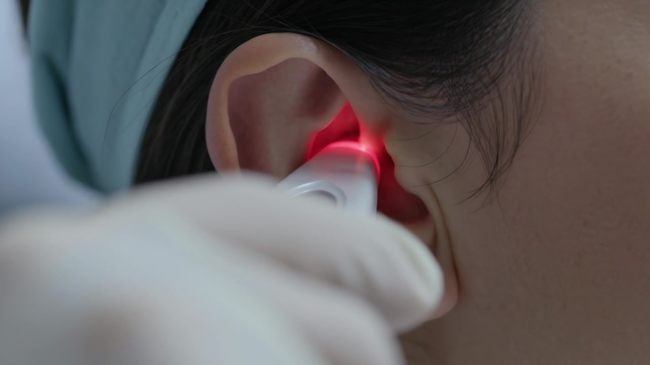 Esaminando un paziente con una sciarpa blu in clinica, una mano guantata inserisce un otoscopio bianco, accendendo un LED rosso.