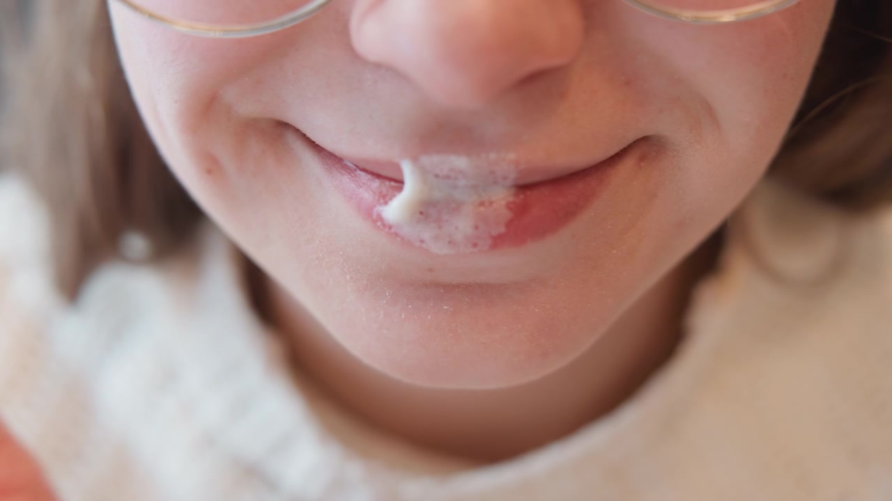 Gros plan intime d'une jeune femme au sourire doux appliquant délicatement du baume à lèvres blanc du bout du doigt, laissant le produit étalé sur les lèvres supérieures et inférieures sous une lumière intérieure diffuse, créant une ambiance domestique calme