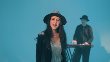 lady in a black hat and leather jacket singing with expressive gestures, her hands and body in motion. a keyboardist is seen in the background