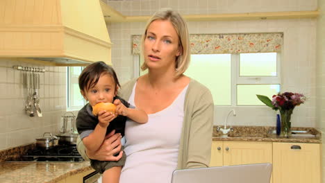tired young mother in kitchen trying to work on laptop holding baby