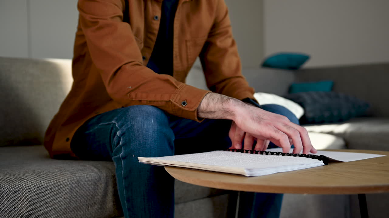 Close Up Of An Unrecognizable Blind Man Reading A Braille Book While Sitting On Sofa At Home