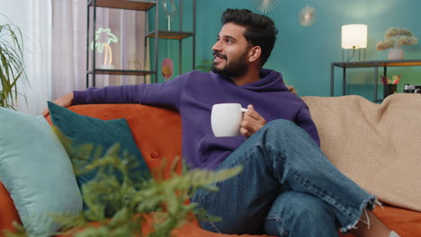 Handsome-smiling-indian-man-drinking-a-cup-of-coffee-or-herbal-tea-sitting-at-home-couch,-break-time