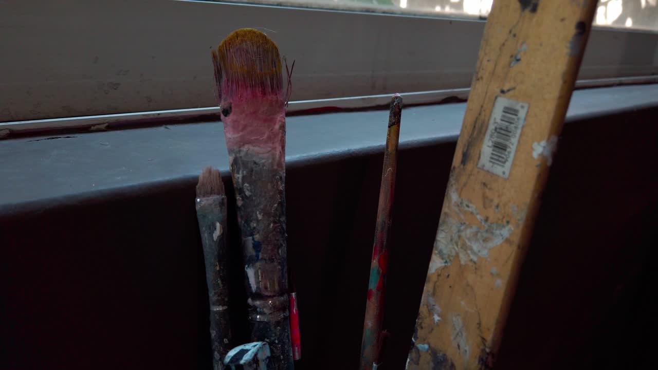 Paint-splattered, used paintbrushes in close-up near a window, with natural daylight and outdoor plants in the background. Slow motion.