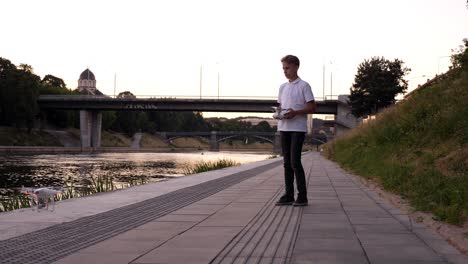 concentrated teenager trying to land the drone near the river, evening long shot