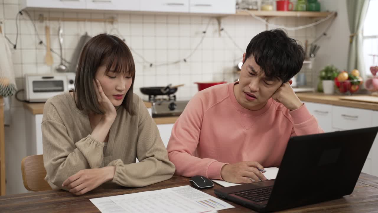 frustrated Asian couple managing finances for the tax season with laptop in the dining room at home. they prop head feeling worried about financial problems while looking at document from bank