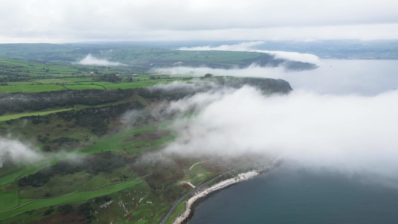 tomada aérea al revés de la hermosa naturaleza de irlanda del norte con vista de la carretera costera cerca de la ciudad de glenarm, rocas naturales y el mar azul y tranquilo por encima de las nubes