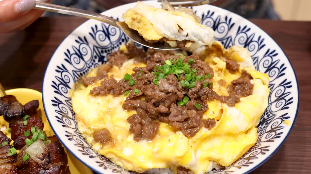 A close-up view of a person using a spoon and fork to scoop a beef and egg rice meal in a brightly lit Bangkok restaurant