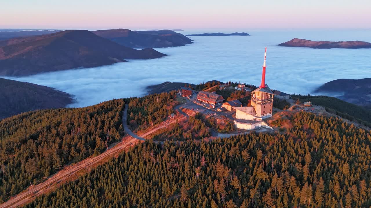 Aerial hyperlapse of Lysá hora at sunrise, telecommunications tower, buildings, and people enjoying the view above a sea of clouds with orange-pink skies. Location: Lysá hora, Czech Republic