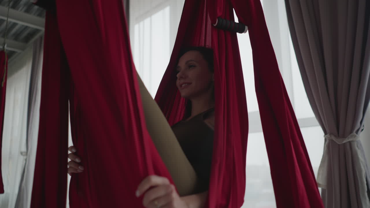Bright yoga studio with floor to ceiling windows and sheer curtains as practitioner wrapped in red silk hammock swings gently smiling, conveying serene balance wellness flow practice
