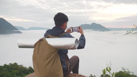 asian hiker male sitting on the rock and using smartphone taking photo of the top view foggy mountain