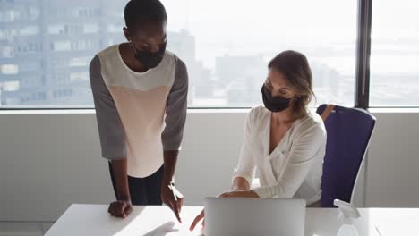 two diverse female colleagues wearing face masks looking at laptop and discussing in office
