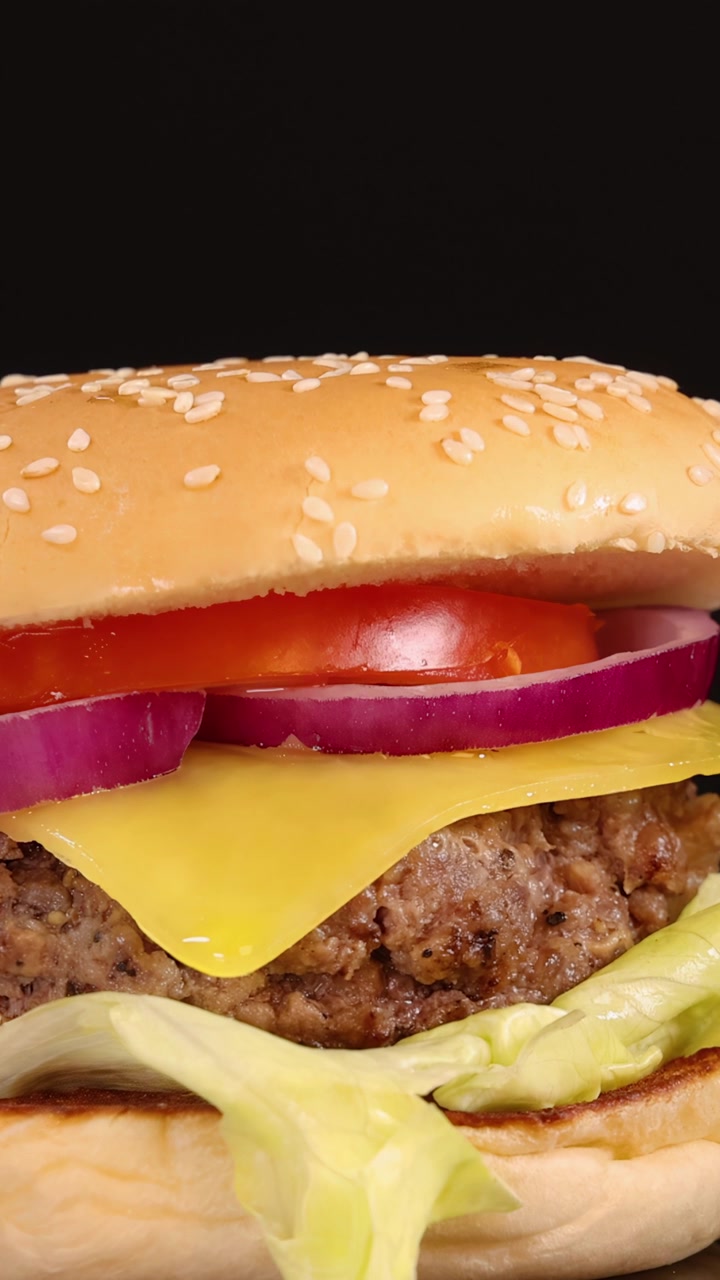 Un succulento cheeseburger con lattuga, pomodoro, cipolla e formaggio cheddar viene lentamente ingrandito, sotto una luminosa illuminazione da studio e uno sfondo nero senza soluzione di continuità