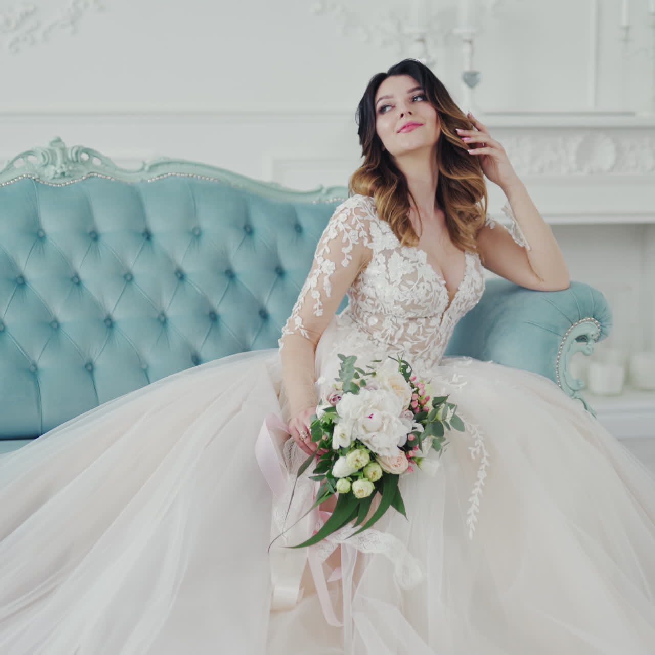 Incredible young woman in a beautiful white wedding dress is sitting on the couch. Lovely model with delicate makeup posing on royal sofa and smiling to camera.