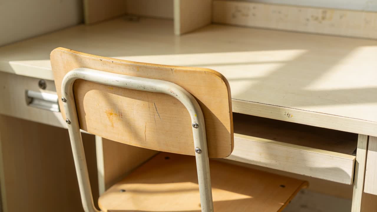 Kamera přibližuje a zaostřuje na dřevěnou židli a stůl školního typu ve studijním koutku, zvýrazňuje škrábance.